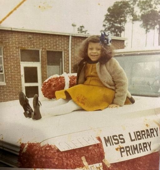 a young girl smiles at the camera while sitting on top of a car that says Miss Library Primary