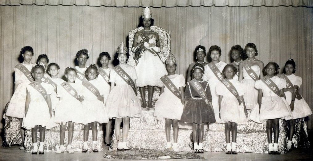 young girls wearing sashes next to another girl with a crown sitting on a chair