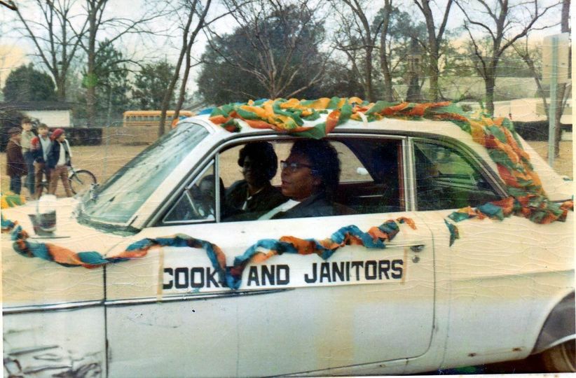 two women ride in a car that says Cooks and Janitors on it