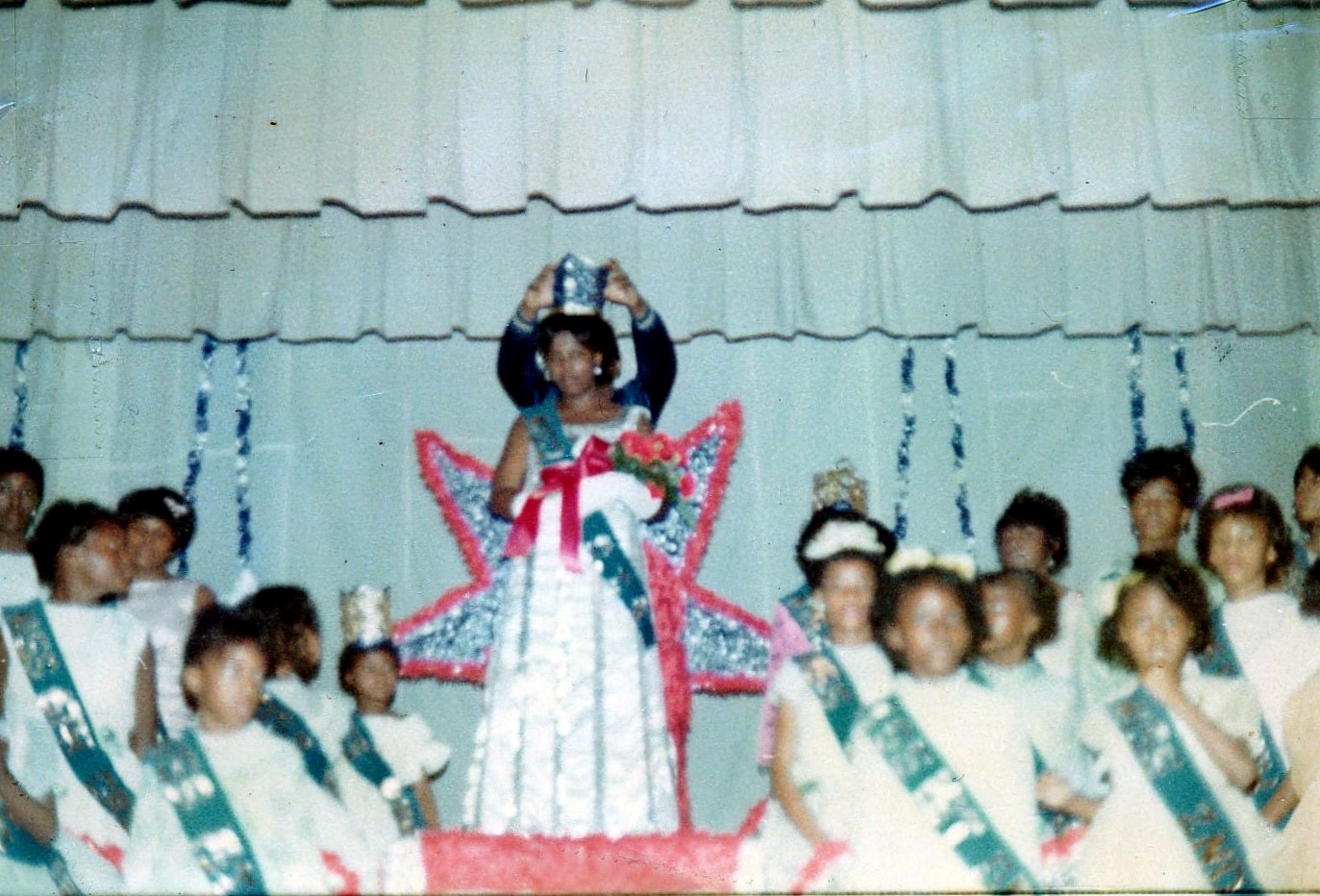 a crown is being placed on a girls head, who is wearing a fancy dress and a sash