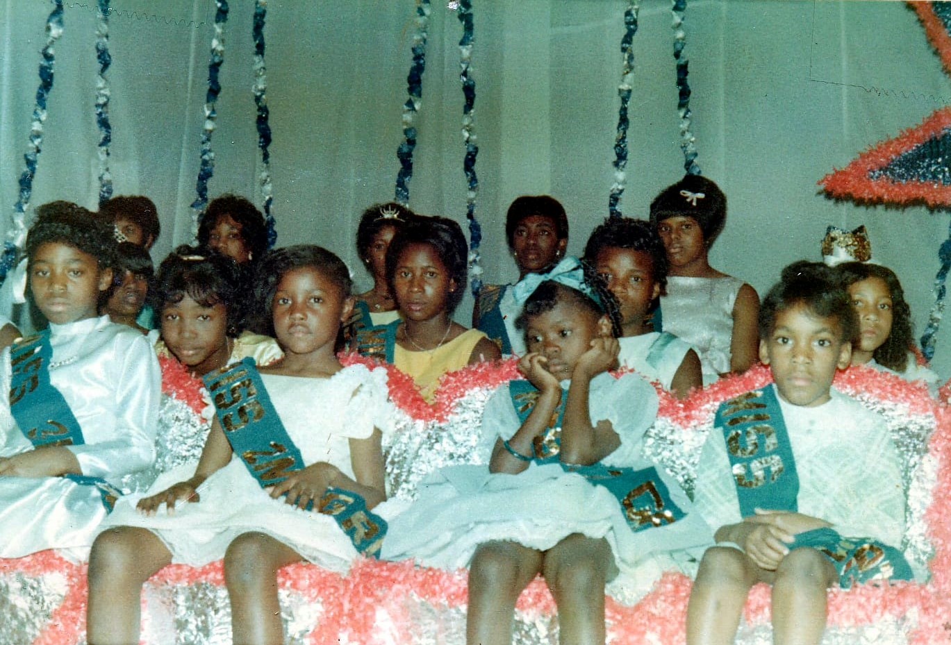 young girls wearing dresses and sashes look tiredly at the camera