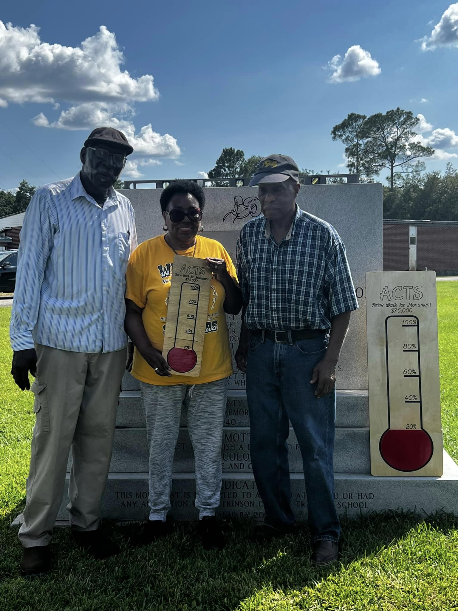 3 people standing in front of the ACTS monument holding a photo that displays the current goal progress